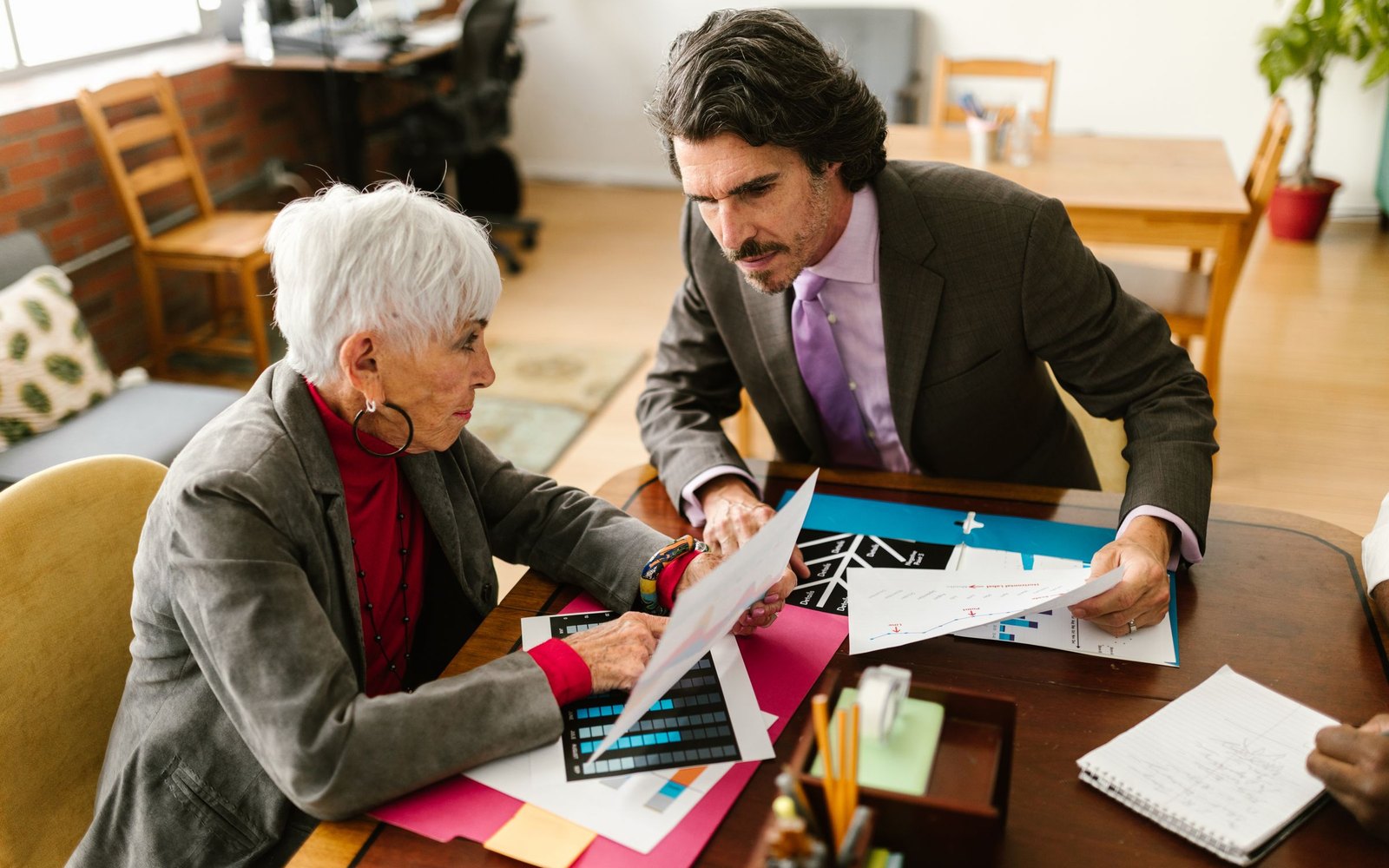 An older woman with short white hair and a man in a gray suit sit at a wooden table, both holding printed documents, with colored charts and a notebook spread between them.