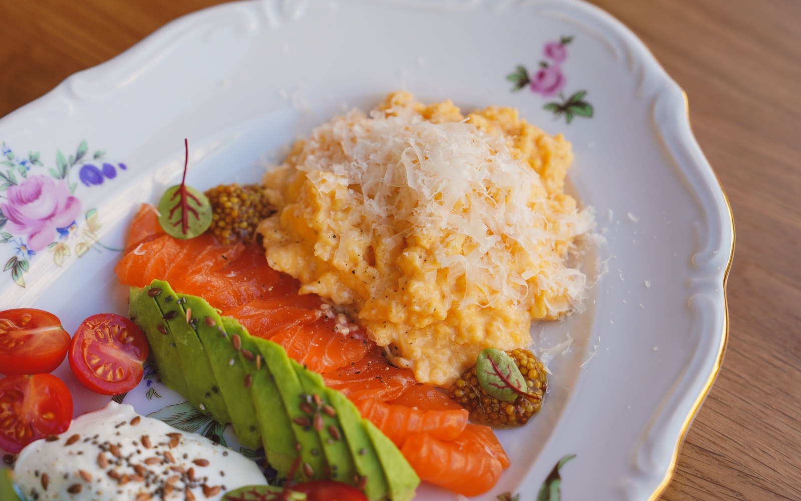 A small floral-rimmed plate holding smoked salmon, sliced avocado, scrambled egg, cherry tomatoes and a spoonful of seeded yogurt.