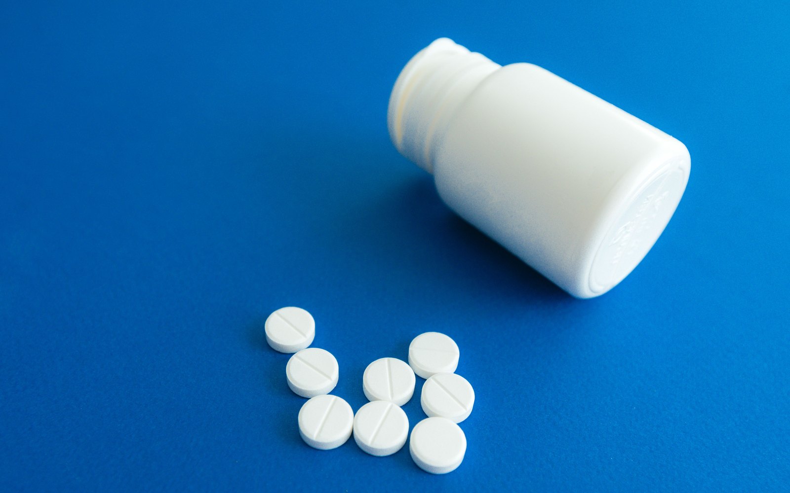 An open white plastic pill bottle on its side beside eight white scored tablets on a blue surface.