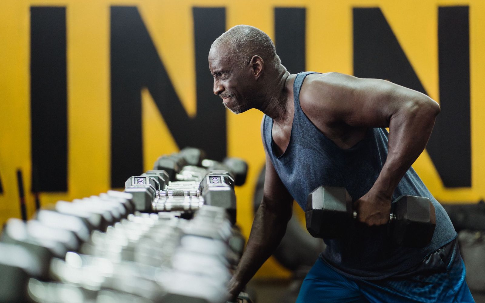 An older Black man performing a one-arm dumbbell row in a gym.