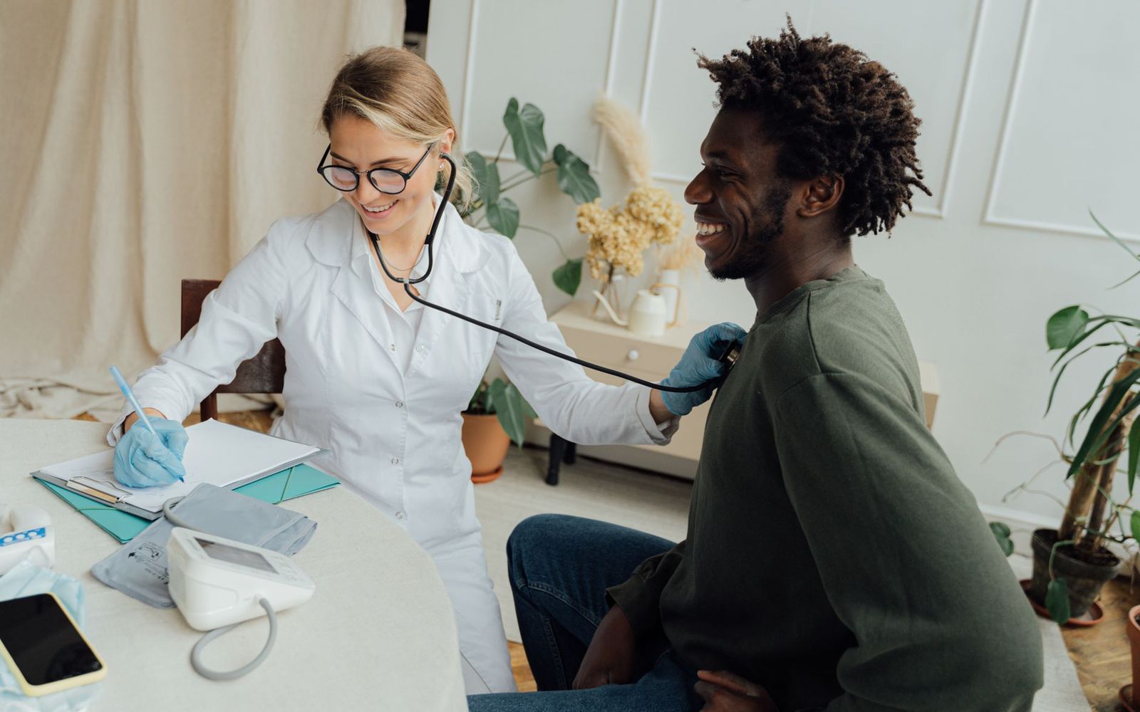 A clinician listening to a smiling man's chest with a stethoscope; a blood pressure cuff sits on the table.