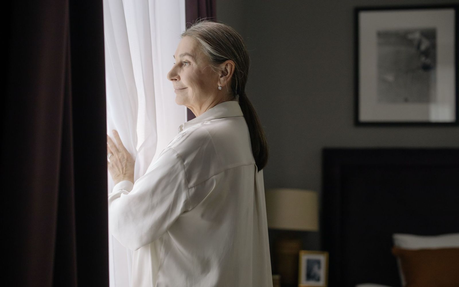A grey-haired woman in a white shirt looking out of a bedroom window in morning light.