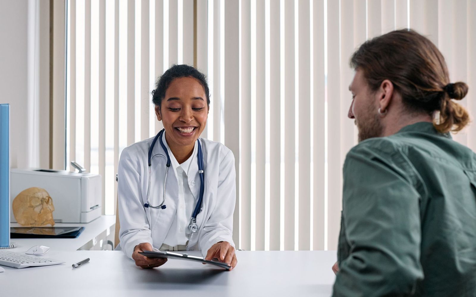 A smiling clinician in a white coat reviewing a clipboard with a patient across a desk.