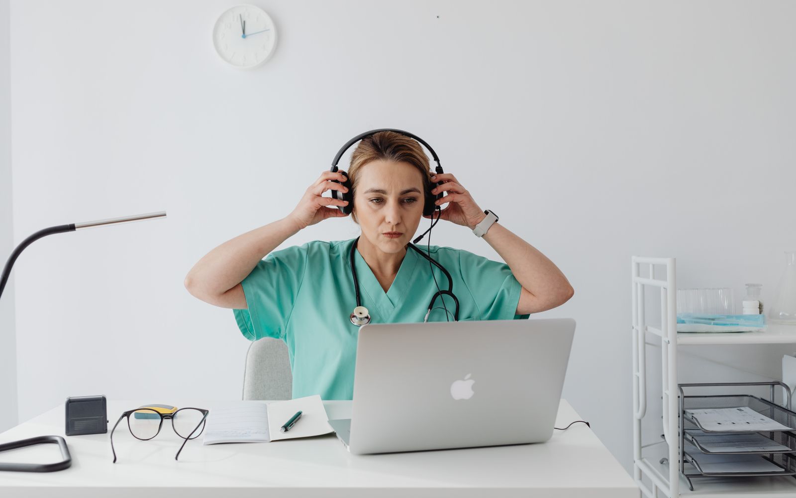 A clinician in teal scrubs with a stethoscope around her neck, putting on a headset at a laptop in a bright clinic room.
