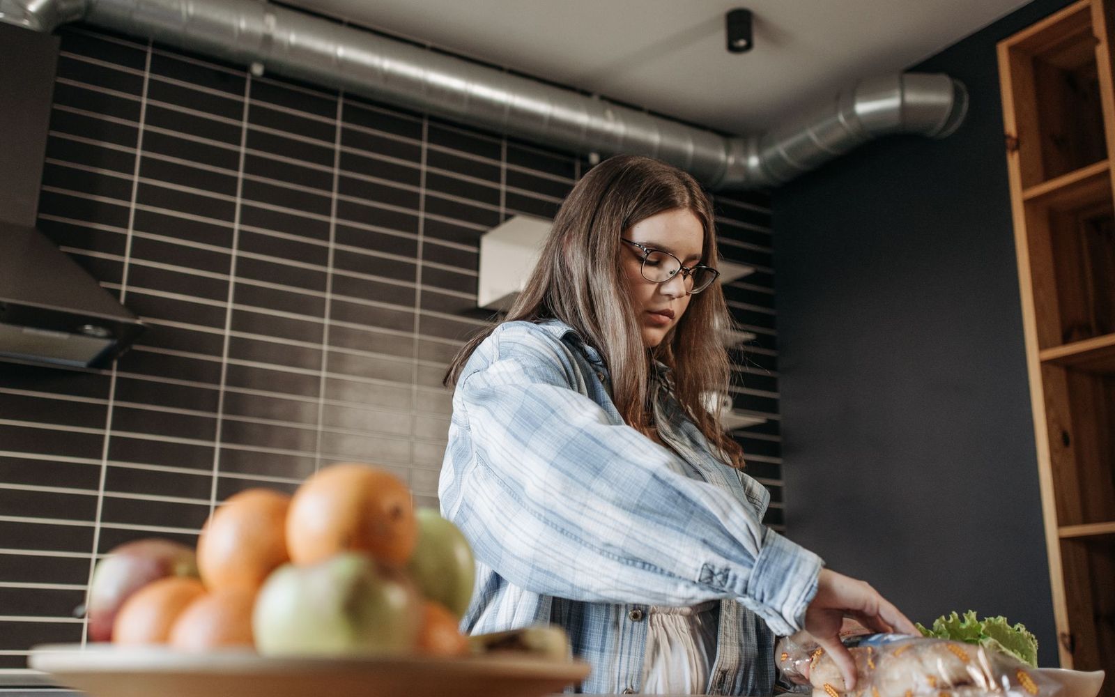 A woman in a plaid shirt preparing vegetables in a kitchen, with a bowl of fruit in the foreground.
