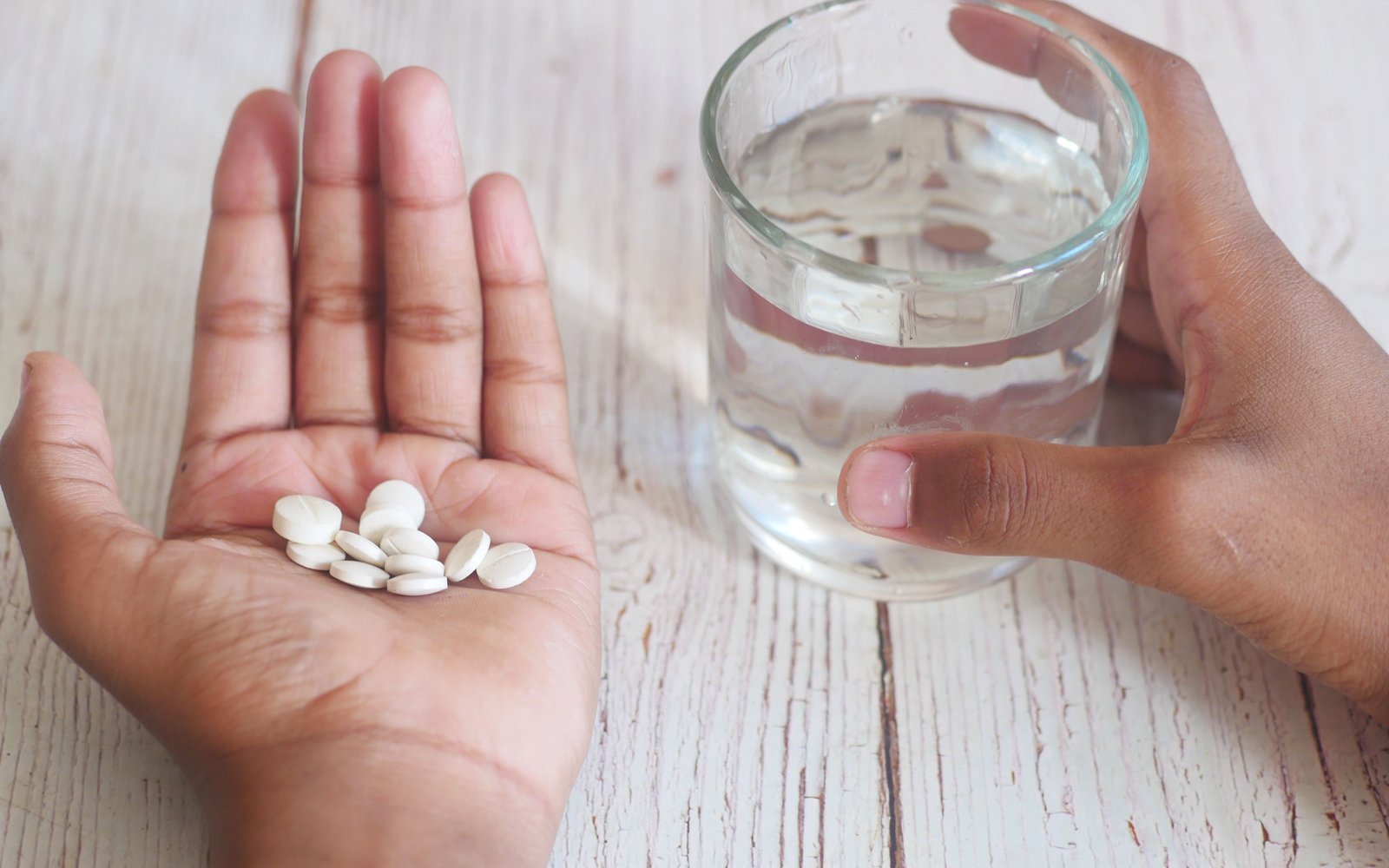 An open palm holding a cluster of small white tablets beside a glass of water steadied by the other hand, on a whitewashed wooden table.