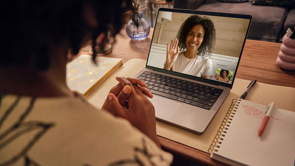 Seen from behind, a person with curly hair sits at a wooden desk with hands clasped, on a laptop video call with a smiling therapist who is waving; a notebook and pen lie beside the laptop.